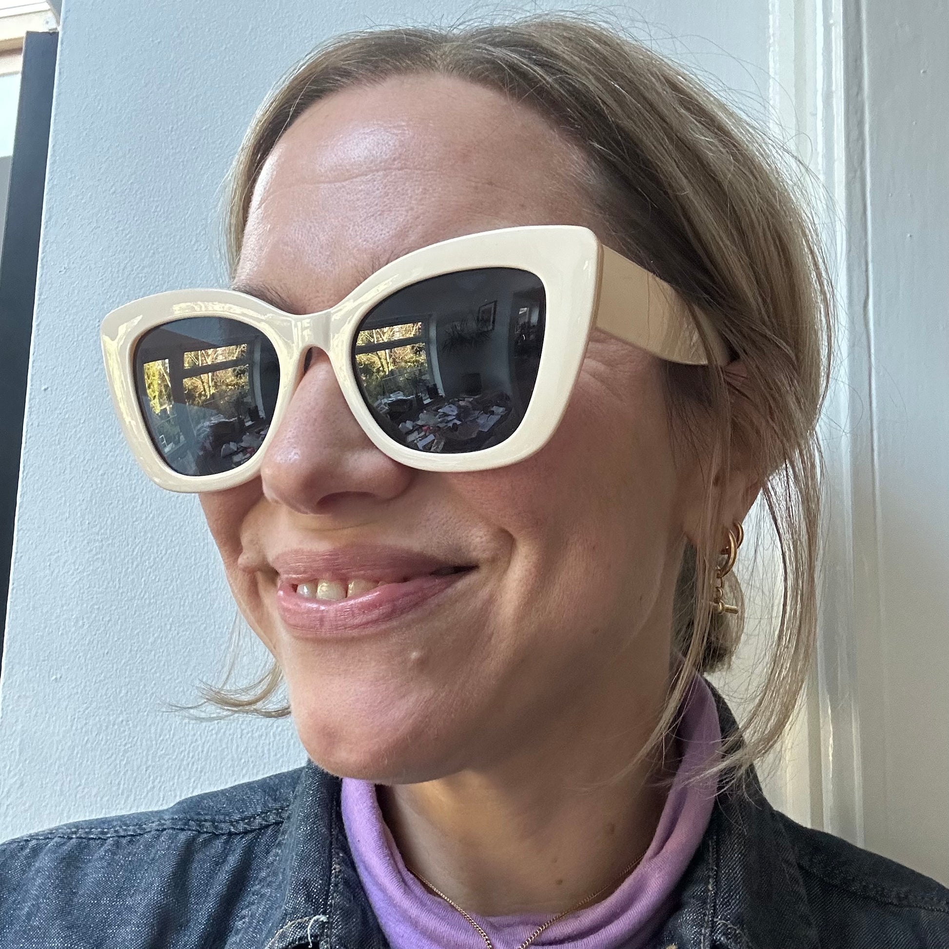 A smiling woman wearing oversized cream-colored sunglasses with dark lenses, styled with a denim jacket, a purple turtleneck, and gold jewelry