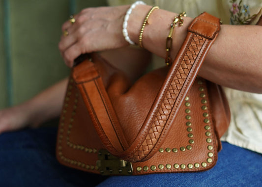 A person's hand holding a cognac-colored vintage style leather handbag with stud detailing and a woven shoulder strap.