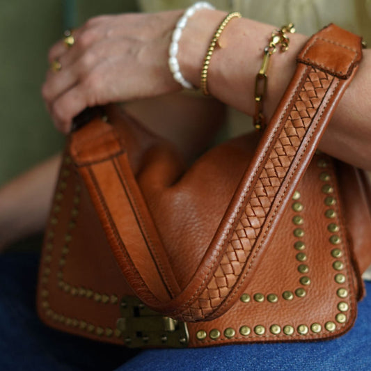 A person's hand holding a cognac-colored vintage style leather handbag with stud detailing and a woven shoulder strap.