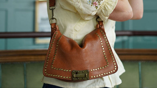 Brown leather handbag with studded details held by a person wearing a white blouse with floral embroidery.