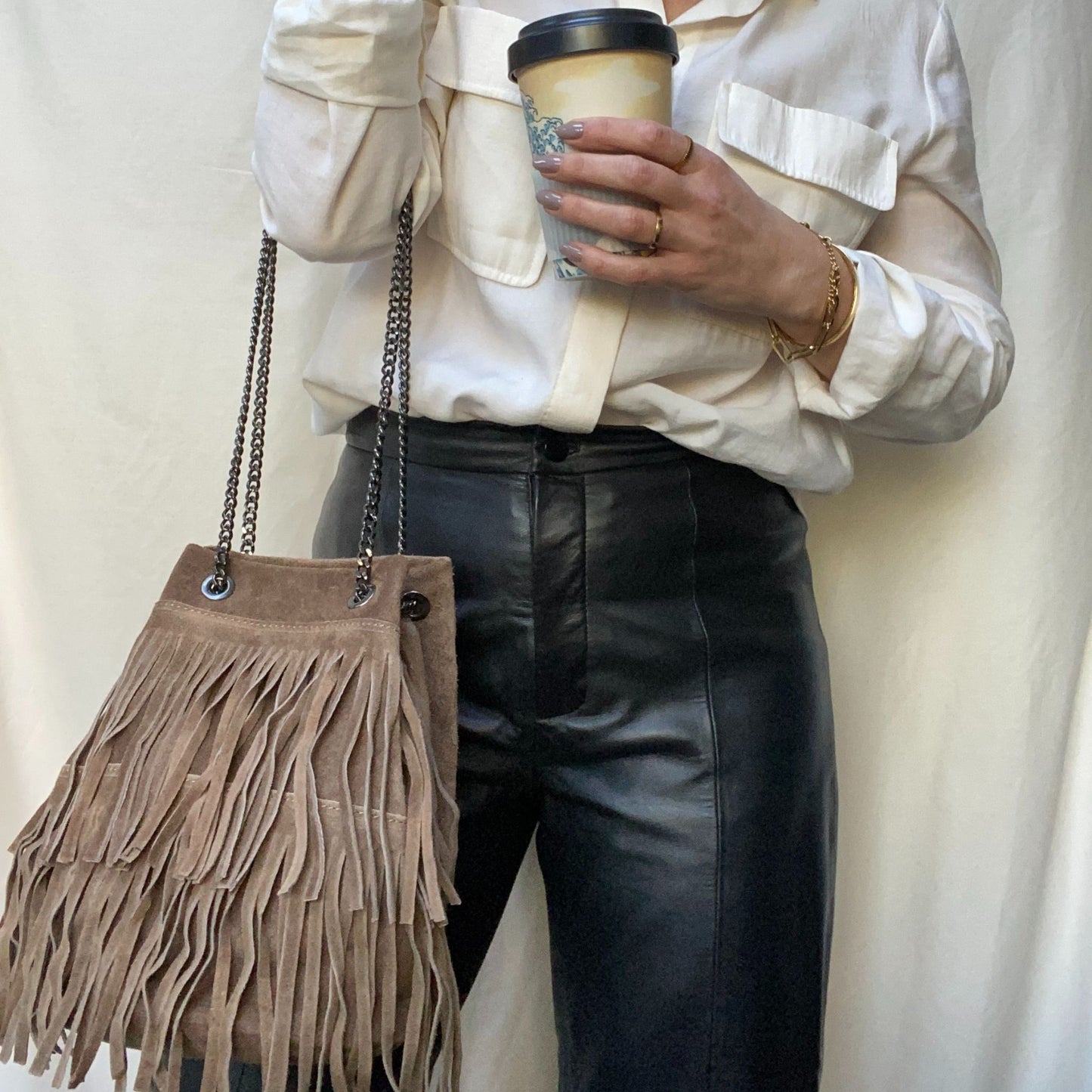 A woman wearing a white shirt and dark pants, holding a taupe fringed suede bag and a cup of coffee.