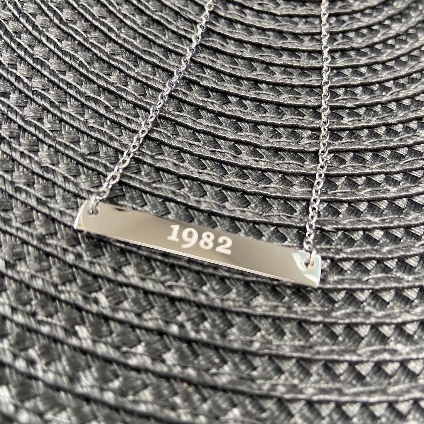 Close-up of a silver bar necklace engraved with '1982' placed on a textured surface