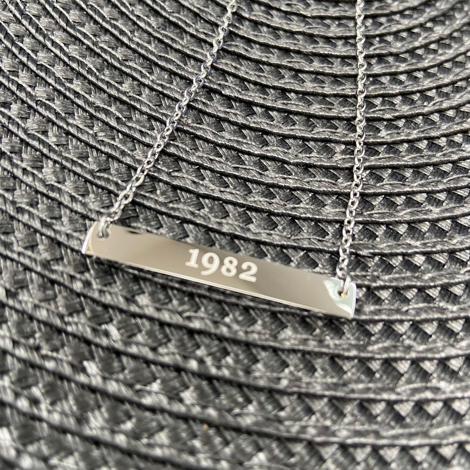 Close-up of a silver bar necklace engraved with '1982' placed on a textured surface