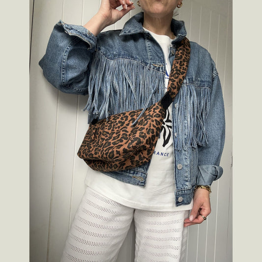 Person wearing a large leopard print cross body sling bag over a fringe denim jacket and white trousers.
